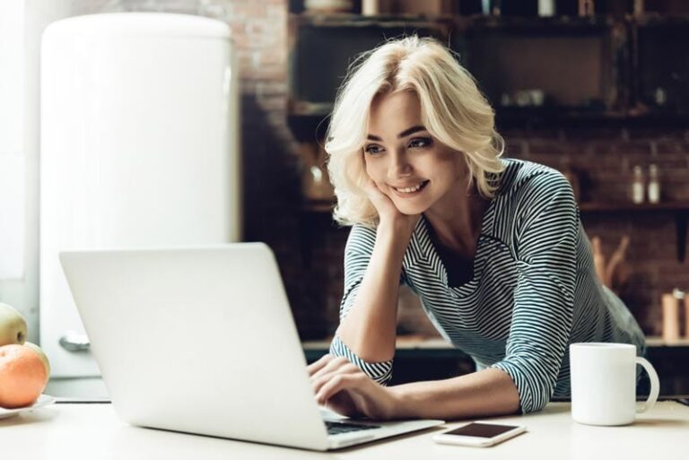 A woman leaning on a kitchen counter, smiling and using a laptop
