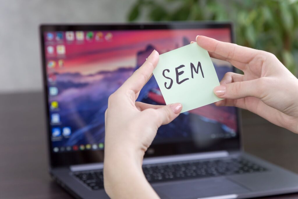 A person's hands holding up a sticky note that says 'SEM' in front of a lap top