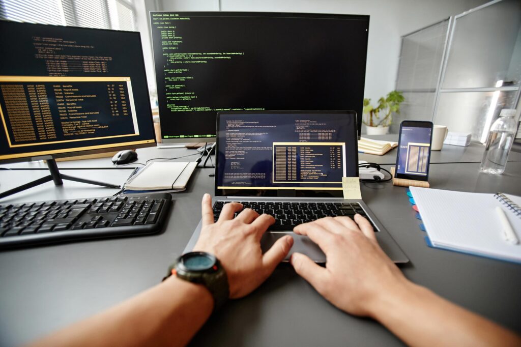 Closeup of person typing code on a laptop with code on monitors behind it